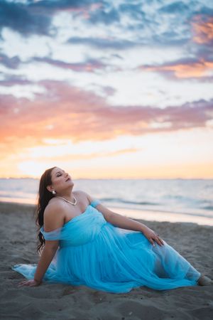 A figure in a flowing turquoise dress poses gracefully on the beach at sunset against a dramatic cloudy sky.