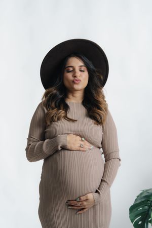 Pregnant woman in beige ribbed sweater and black hat poses with hands on belly against white background.