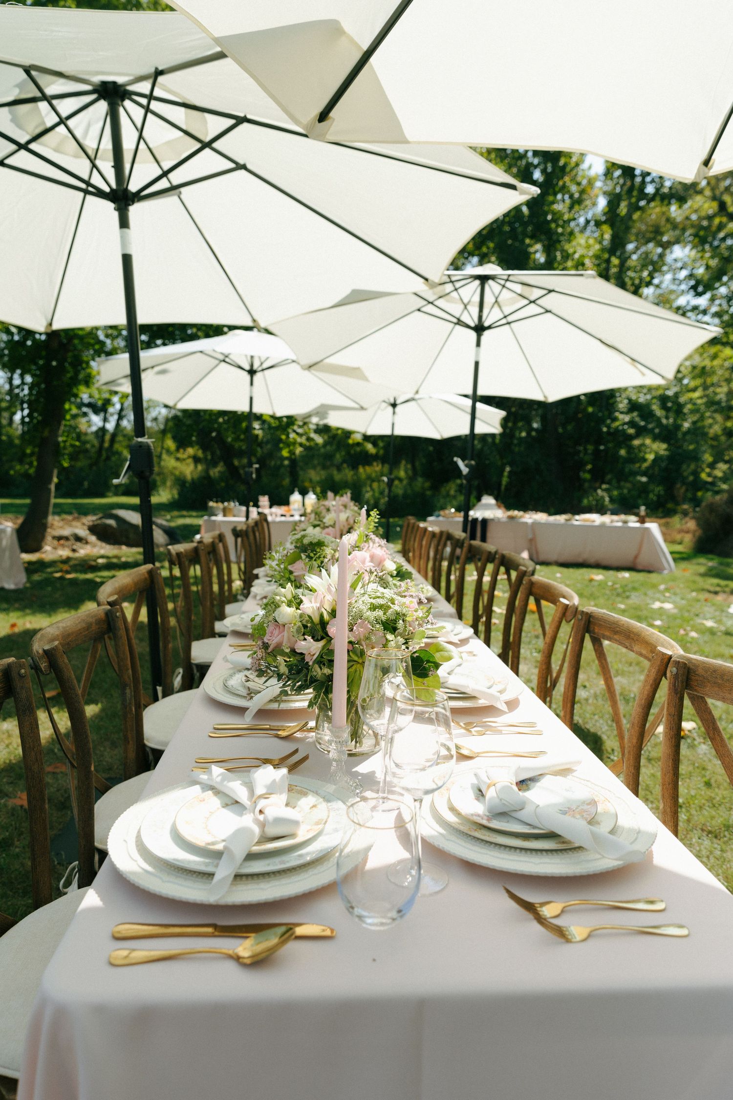 An elegant outdoor dining table set with white linens, gold flatware and umbrellas for a garden wedding reception.