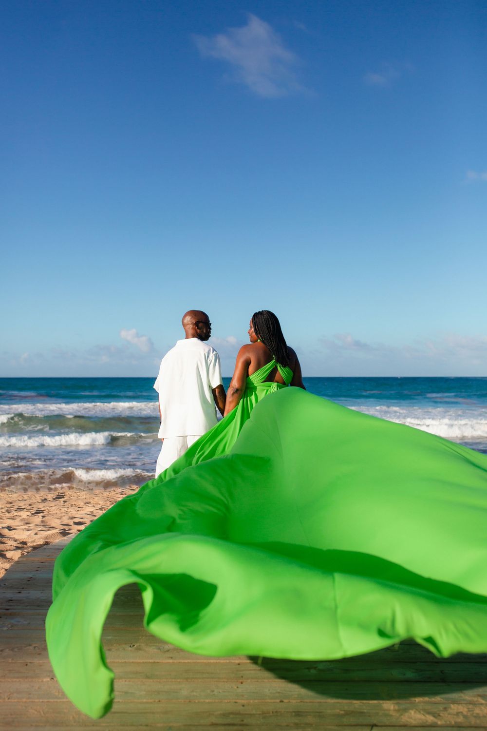 A couple stands on a beach as a vibrant green fabric billows dramatically in the wind against blue ocean waves.