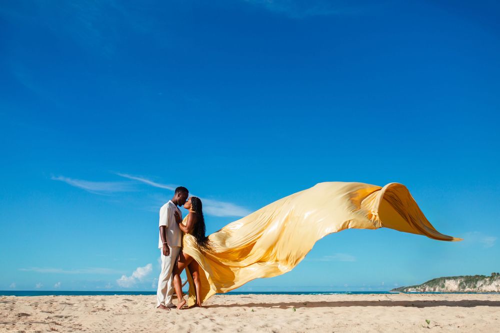 A flowing yellow fabric billows in the wind against a bright blue sky on a sandy beach landscape.