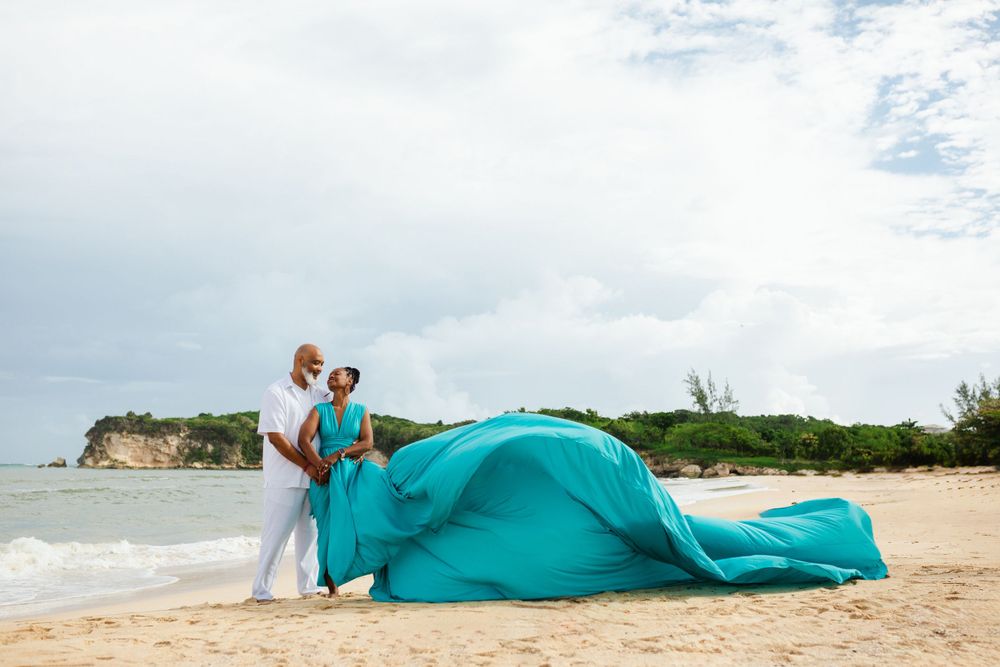 A couple in turquoise attire poses romantically on a sandy beach with cloudy skies in the background.