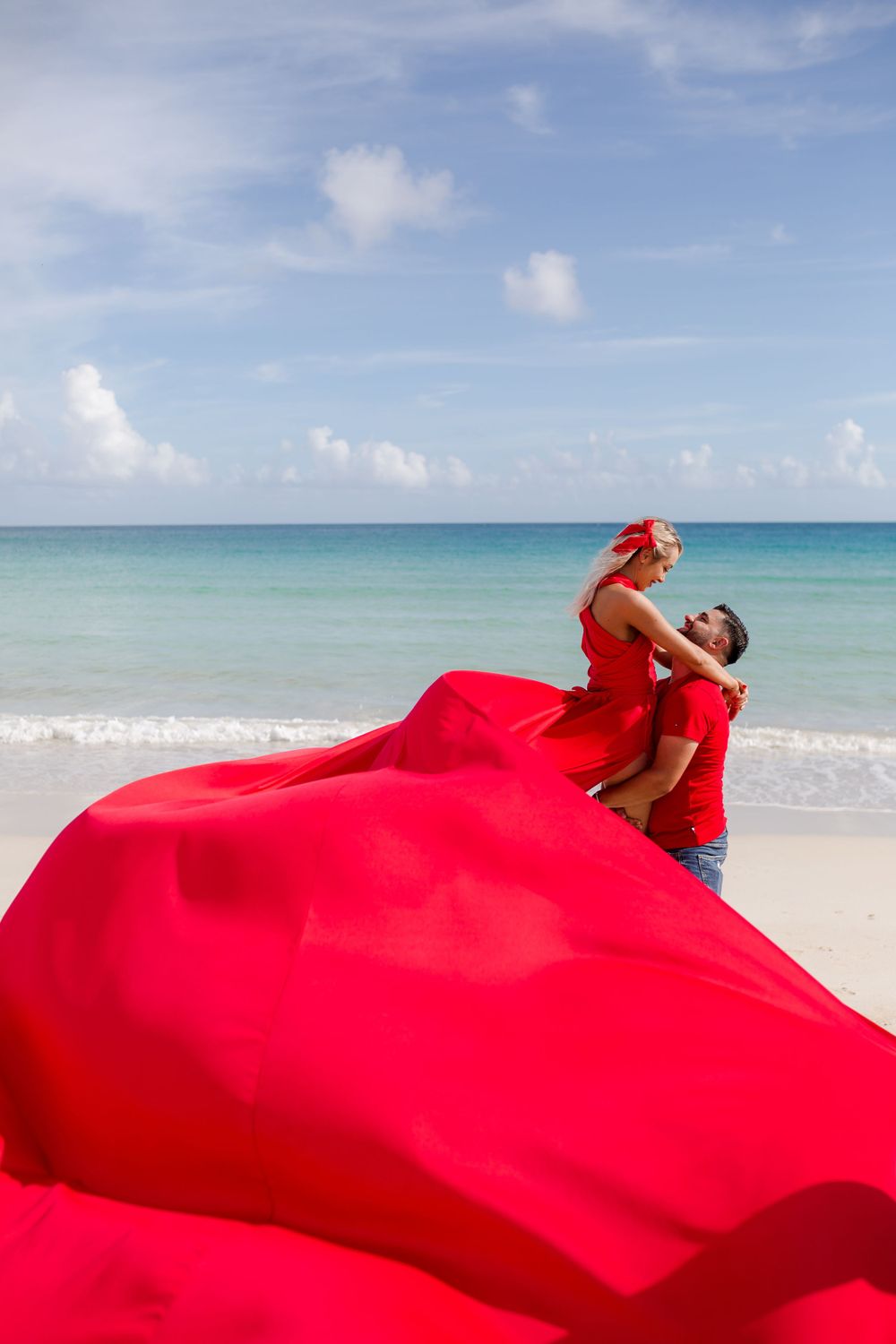 A flowing red fabric billows dramatically against a tropical beach landscape with turquoise water and blue sky.