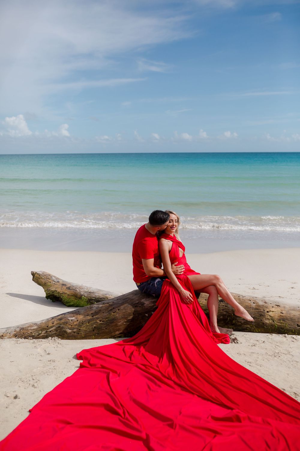 Flowing red fabric dress creates dramatic movement against white sandy beach and turquoise ocean backdrop.