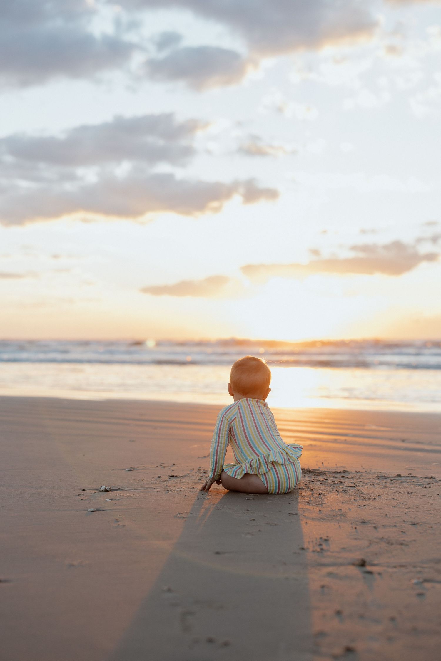 A figure sits alone on a sandy beach at sunset watching the golden sun sink into the ocean horizon.