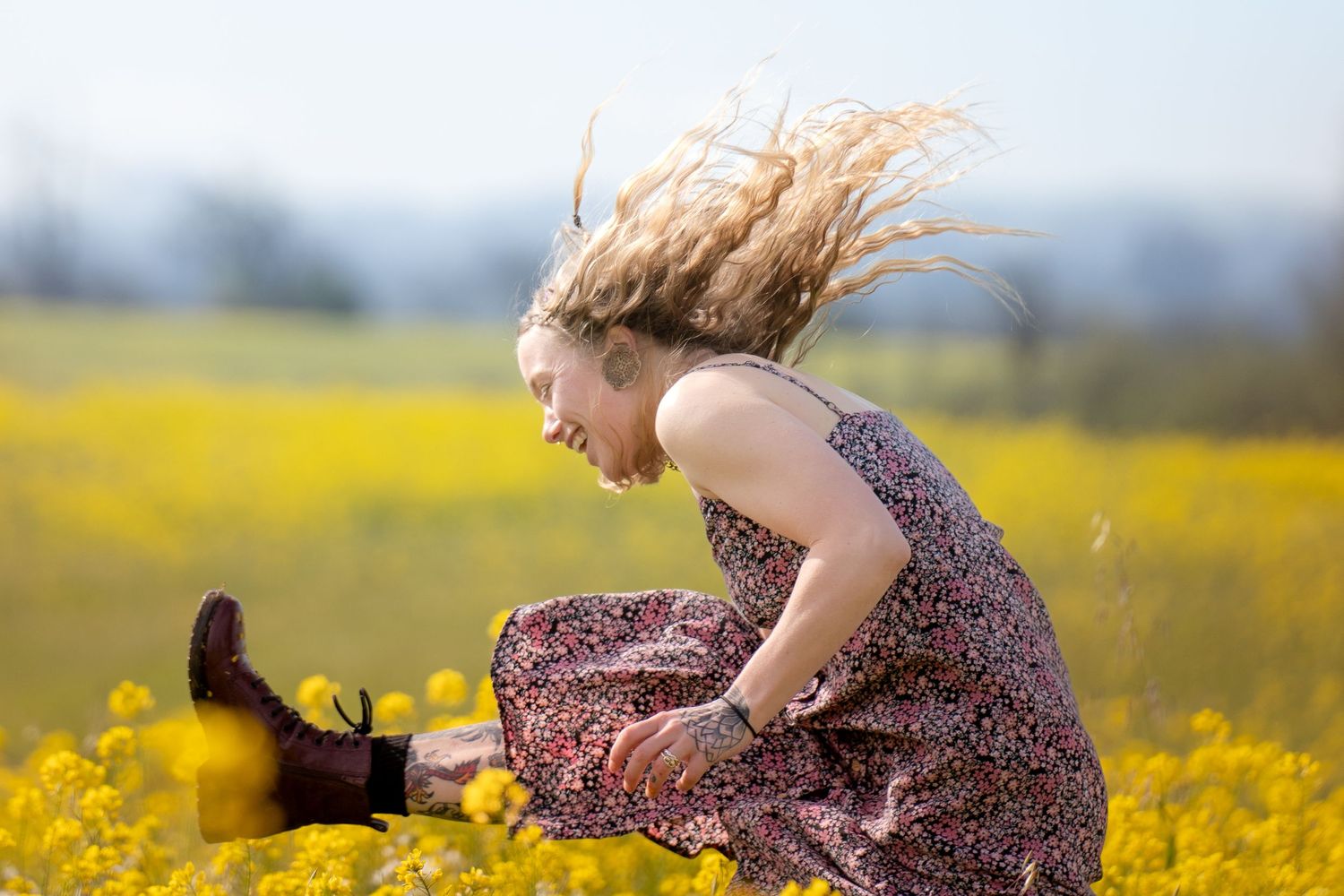 Person in a floral dress with windswept hair jumps in a vibrant yellow flower field.
