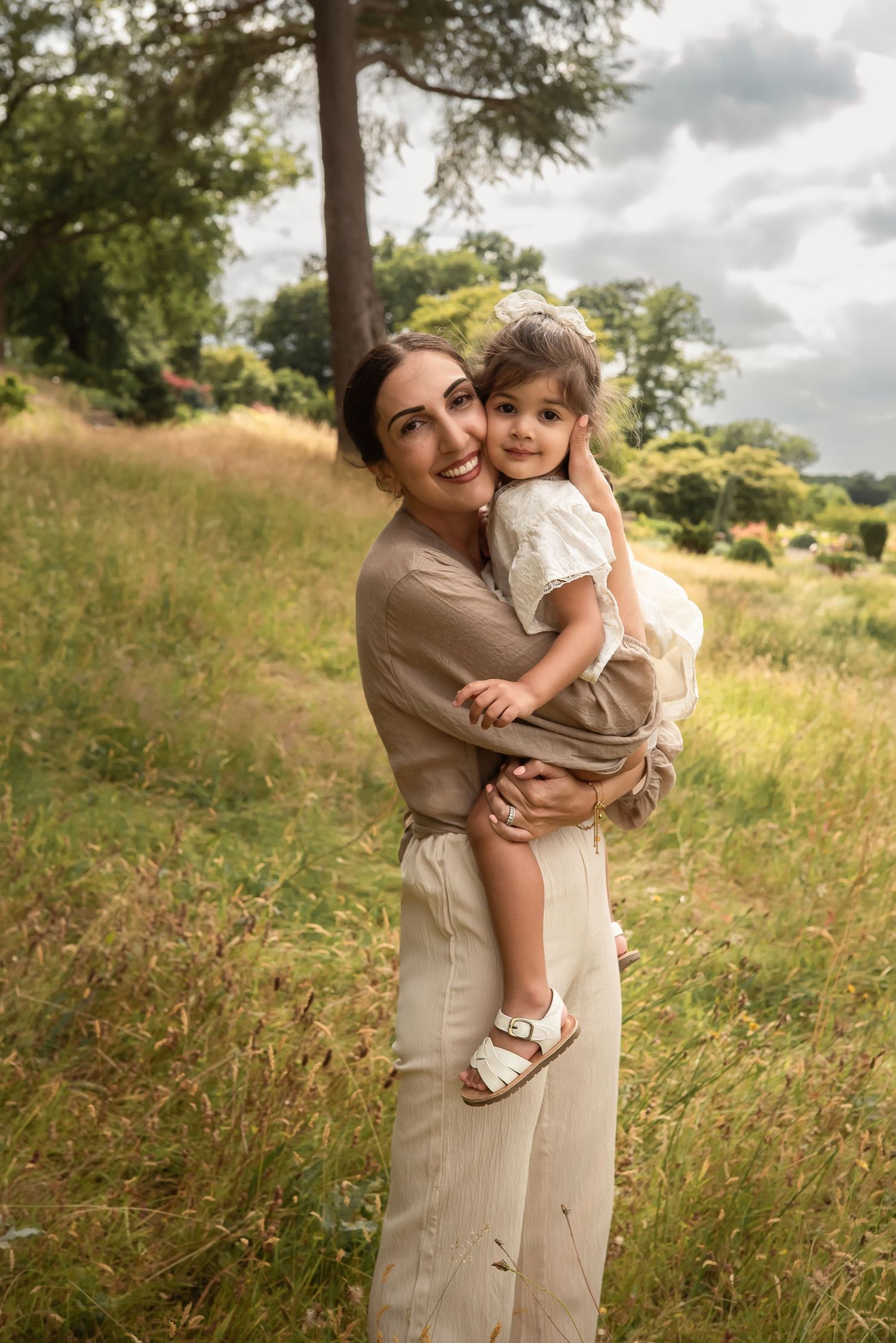 A mother holds her toddler in a sunny meadow, both wearing neutral tones, smiling warmly amid lush greenery.