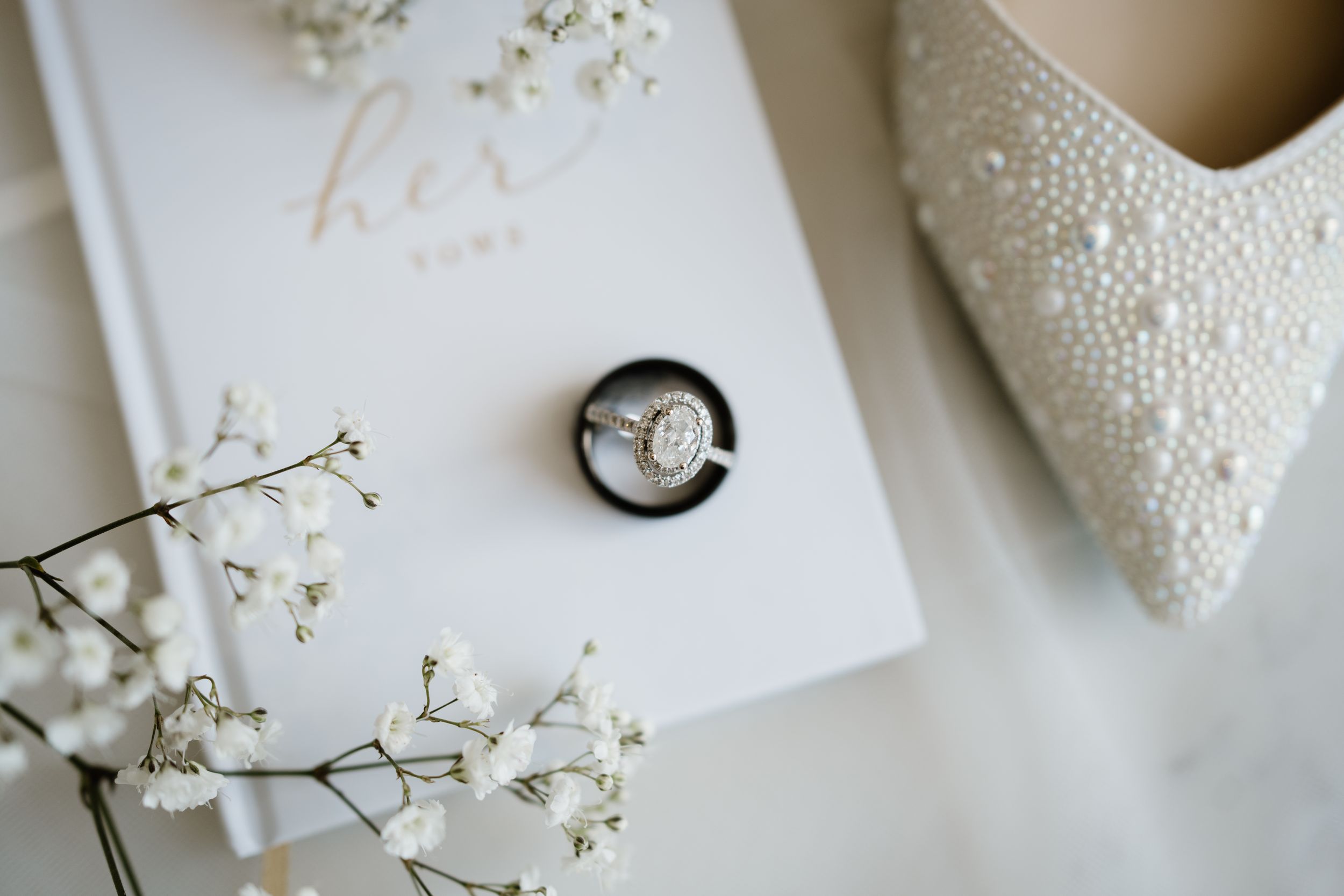 Wedding rings in black box alongside white vows book, delicate baby's breath flowers and bridal heel in soft lighting.