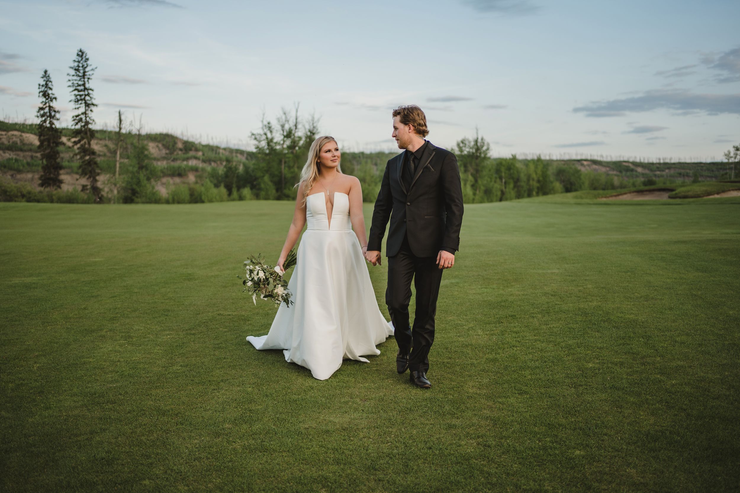 A couple in wedding attire walks hand in hand across an expansive green golf course under a cloudy sky.