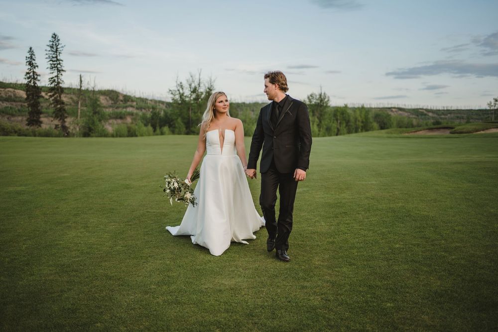 A couple in wedding attire walks hand in hand across an expansive green golf course under a cloudy sky.