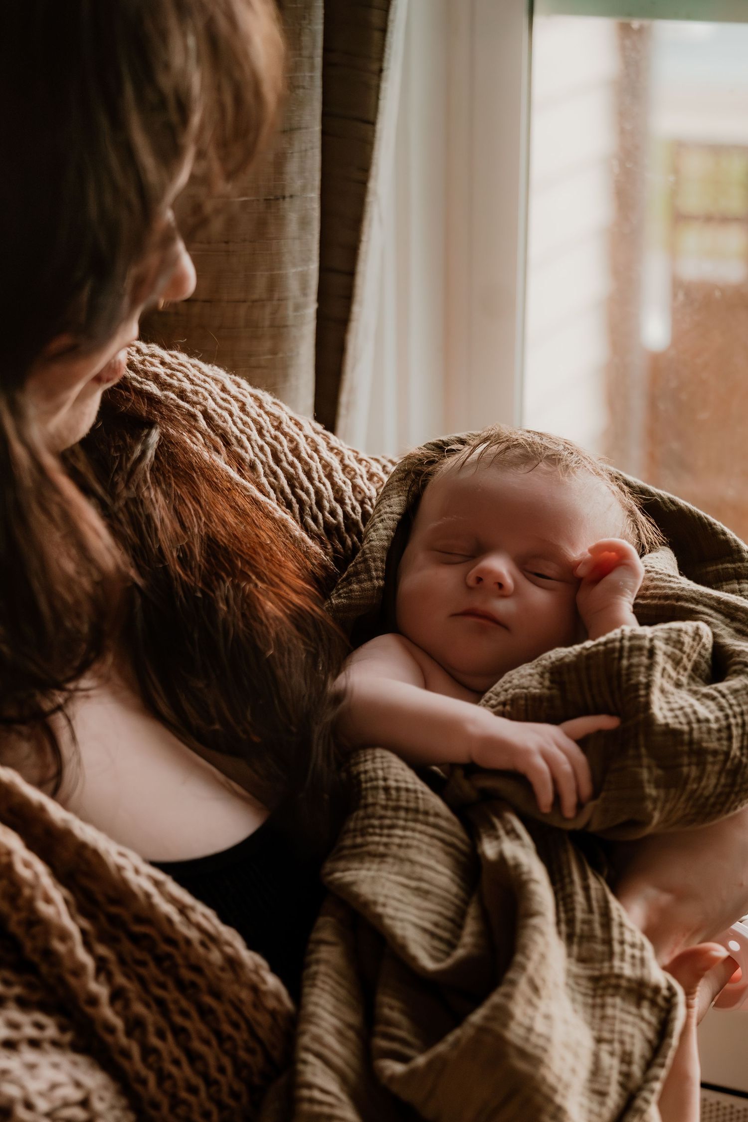 Mother tenderly holding sleeping newborn baby wrapped in olive green blanket near a sunlit window.