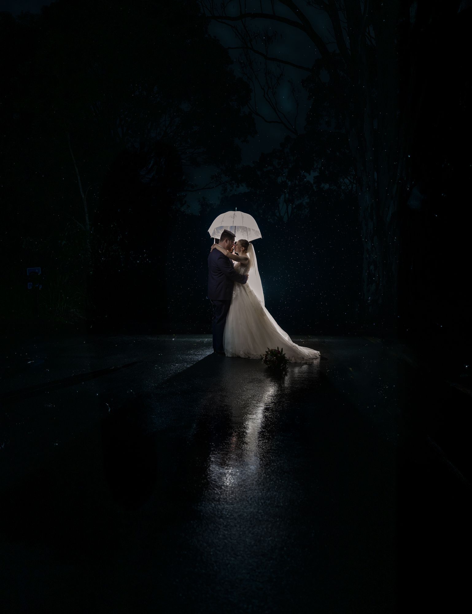 A couple stands under a white umbrella in a dark, moody scene with their reflection visible in still water.