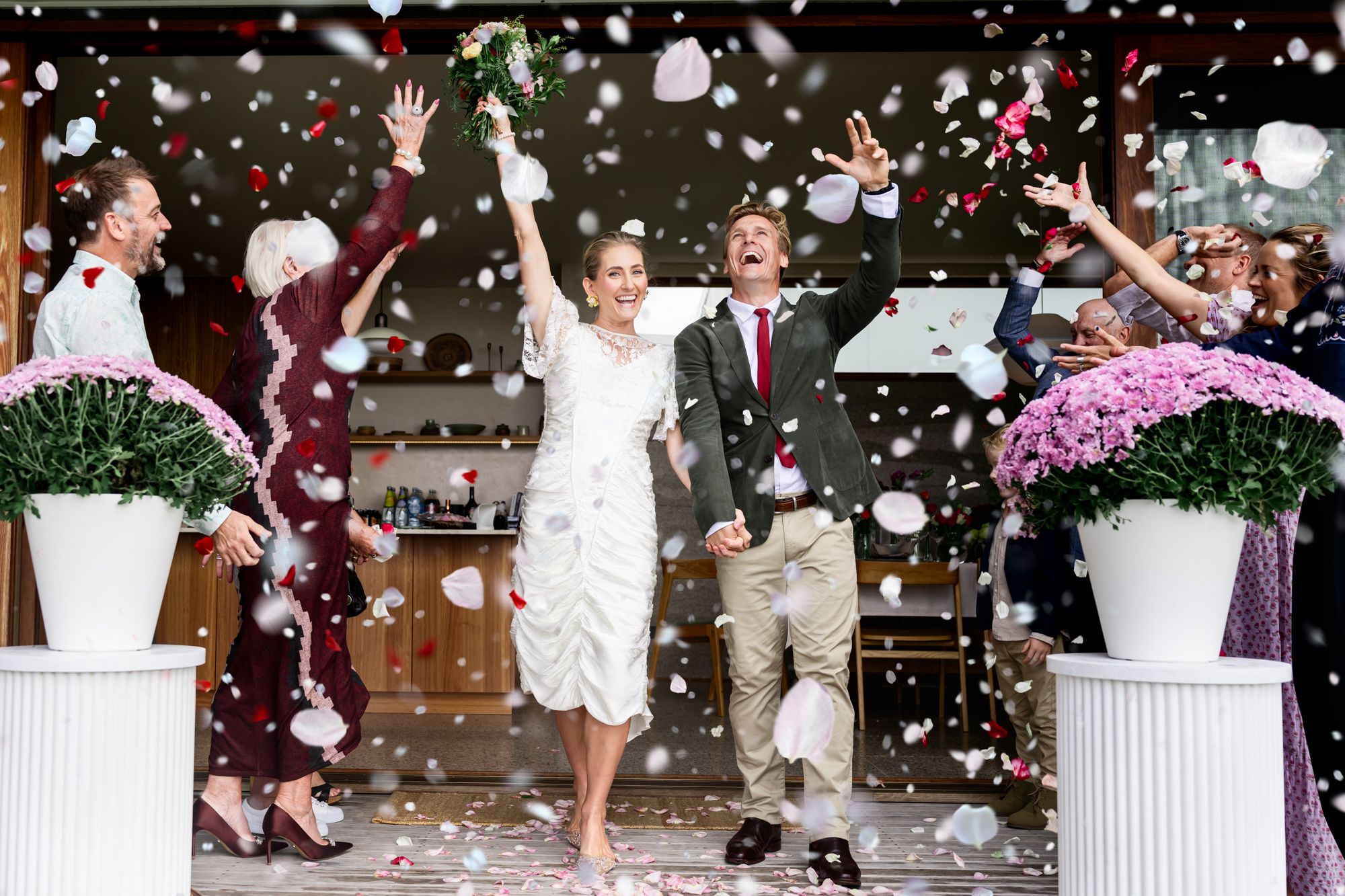Newlyweds exit church celebration through shower of red and white confetti between decorative white floral arrangements.