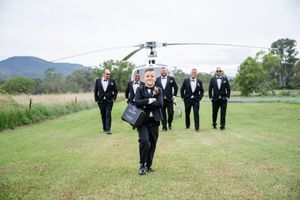 Group in formal attire walking across grass field with helicopter in background.