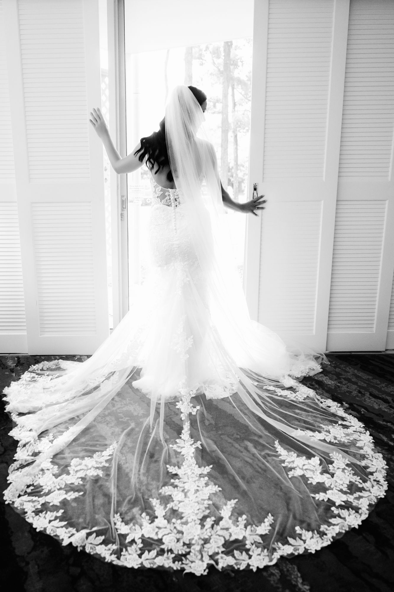 A bride in a flowing lace dress with cathedral veil poses dramatically by a window in black and white photography.