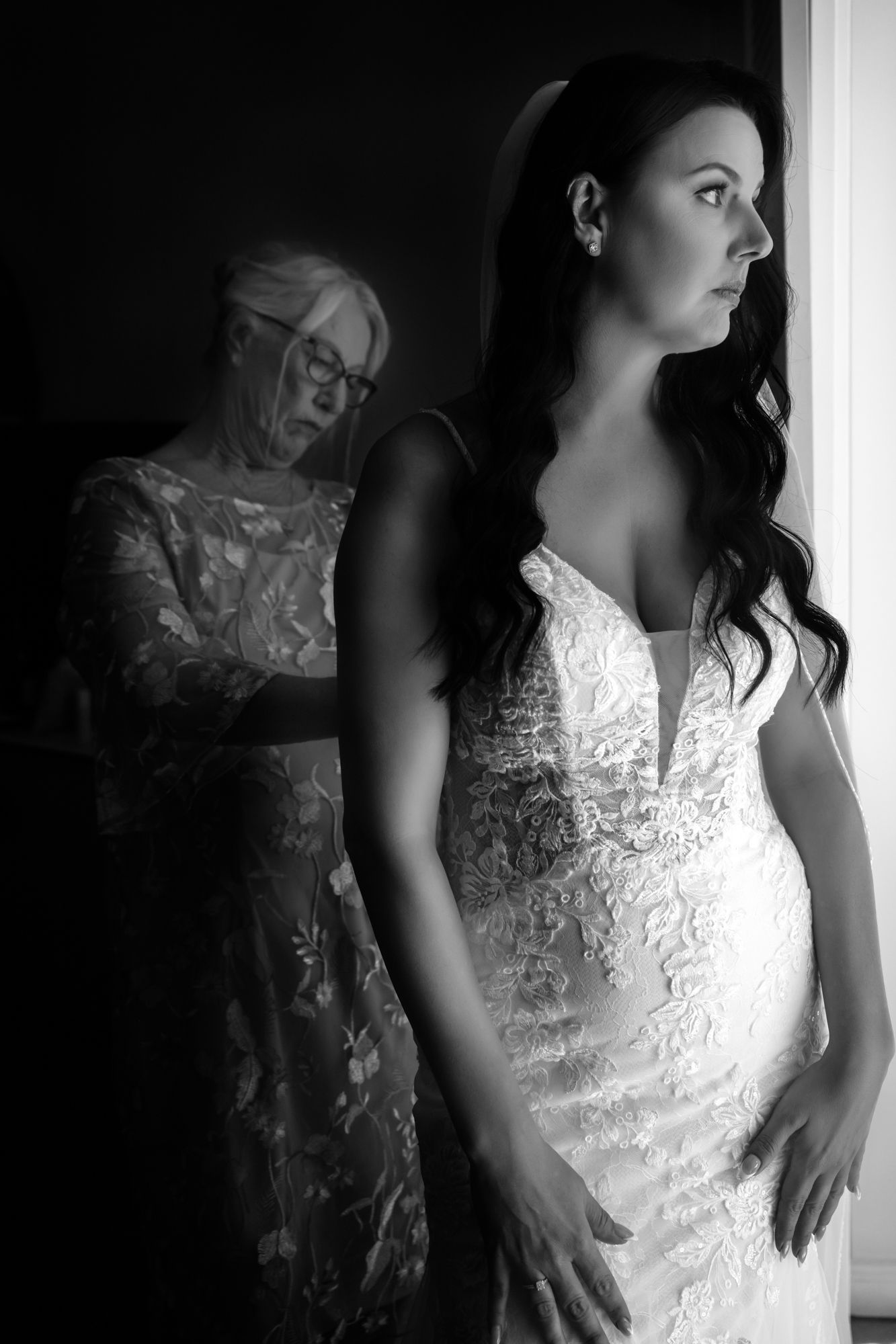 Black and white photograph of a bride in an elegant lace wedding dress standing by a window with soft natural light.