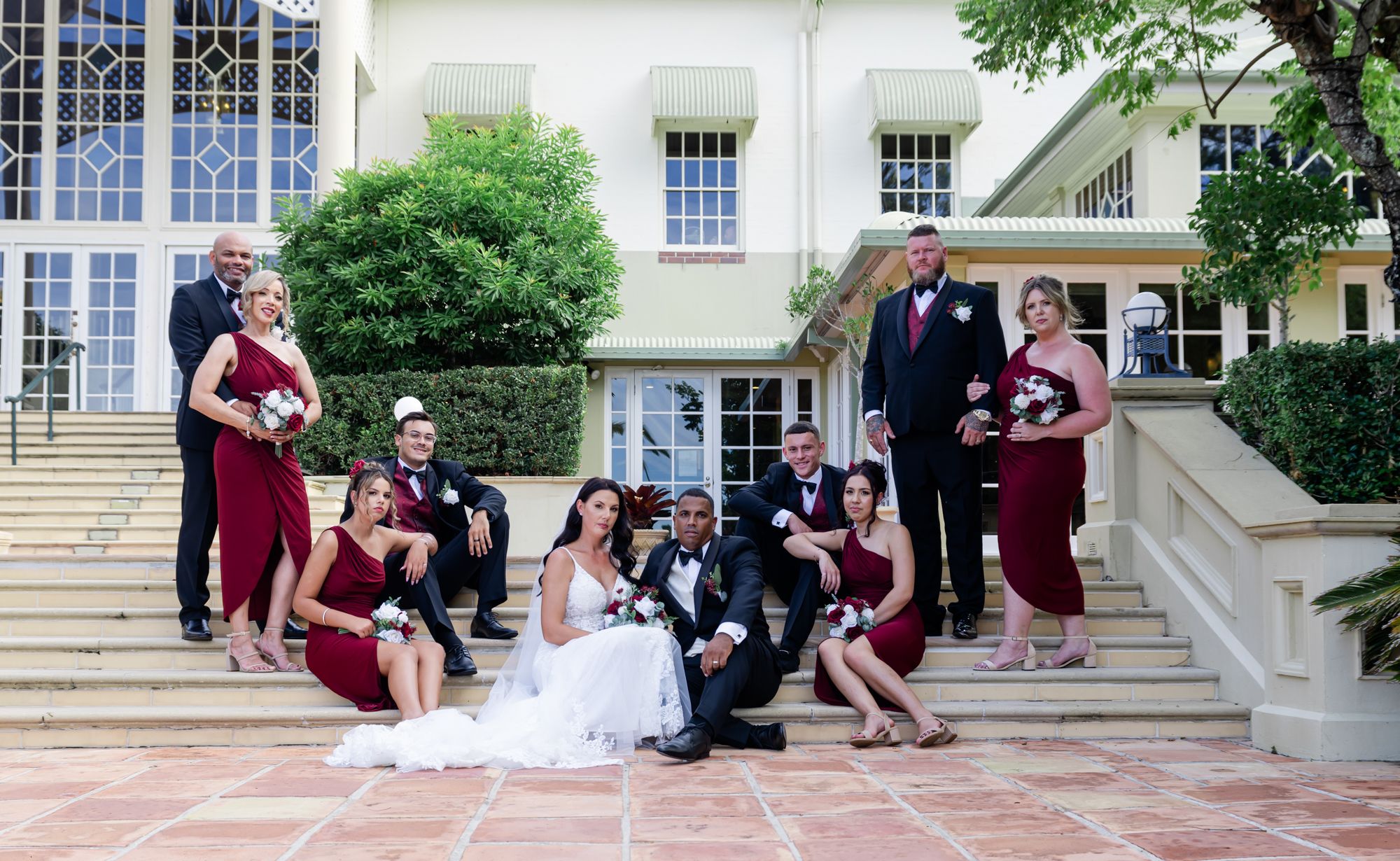 A bridal party in burgundy dresses and black tuxedos poses on stone steps outside an elegant building.