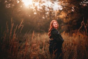 Redhead woman in black jacket standing in golden autumn field at sunset.