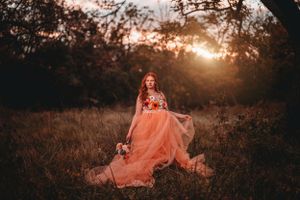 Woman in peach tulle gown holding bouquet in glowing sunset forest clearing.