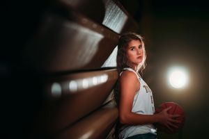 Young woman in white tank top leaning against wall holding red ball in dim light.
