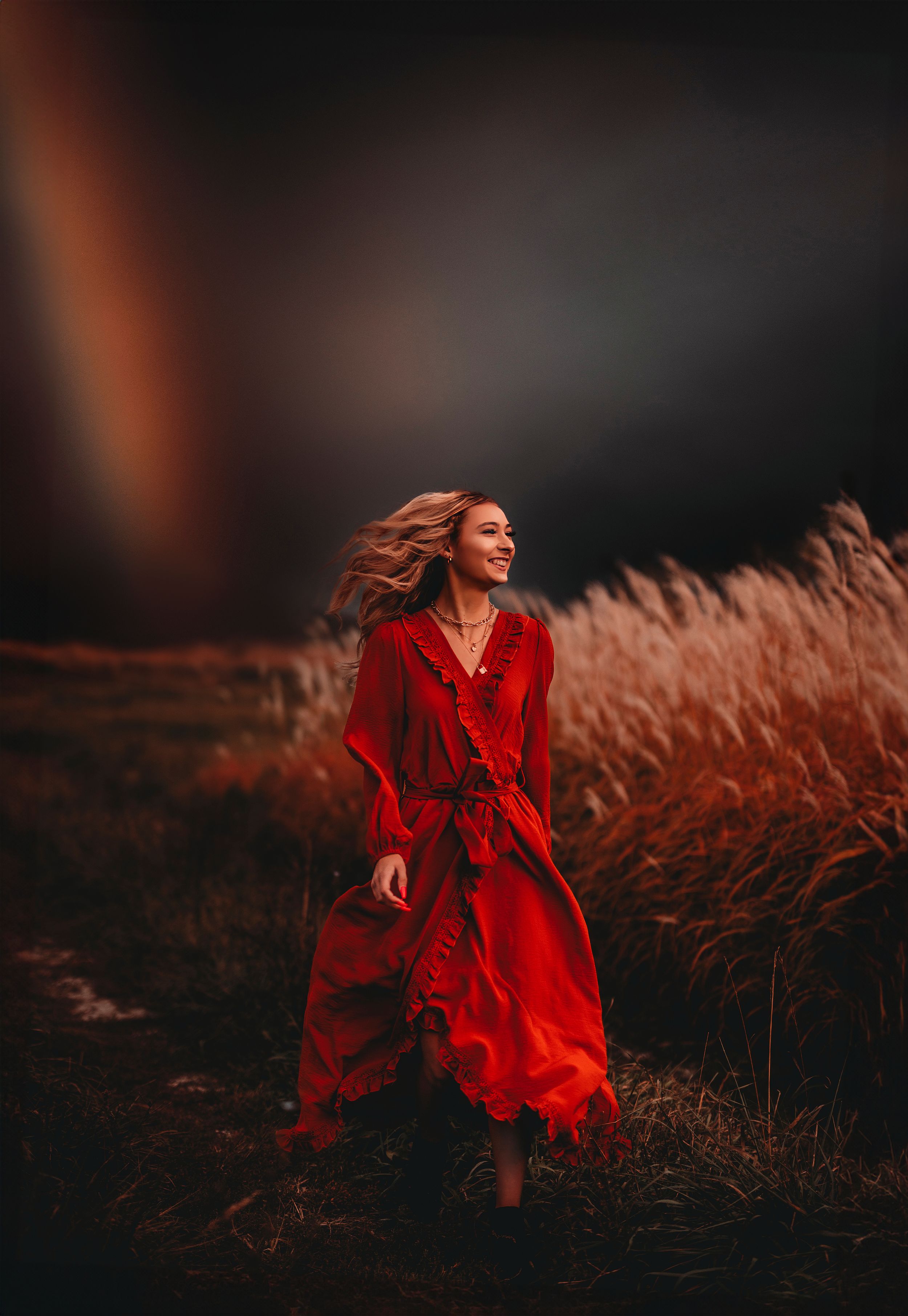 Smiling blonde woman in red dress walking through field with rainbow backdrop.