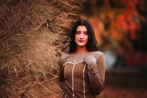 Dark-haired woman in brown sweater posing beside tall dried grass in autumn park.