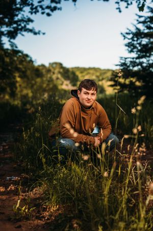 Young man in brown hoodie crouching in tall green grass in a sunlit forest clearing during golden hour.