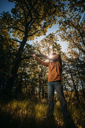 Man in brown hoodie aiming a rifle in a sunlit forest with golden leaves and bright sun behind the trees.