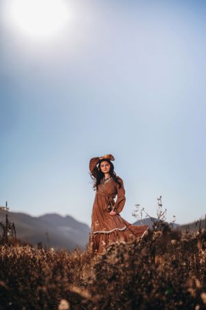 Woman in flowing brown boho dress standing in sunlit golden field with mountains.