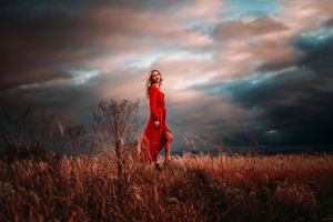 Woman in red dress standing in golden field under dramatic cloudy sky.