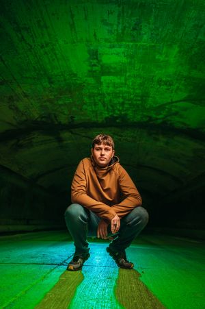 Young man in brown hoodie crouching in dramatic green-lit tunnel space.