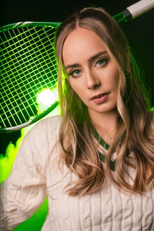 Young woman holding a green badminton racket wearing a white cable-knit sweater with green neon lighting.