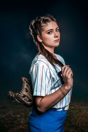 Young woman in baseball uniform holding a glove, posing dramatically against a dark smoky background outdoors.