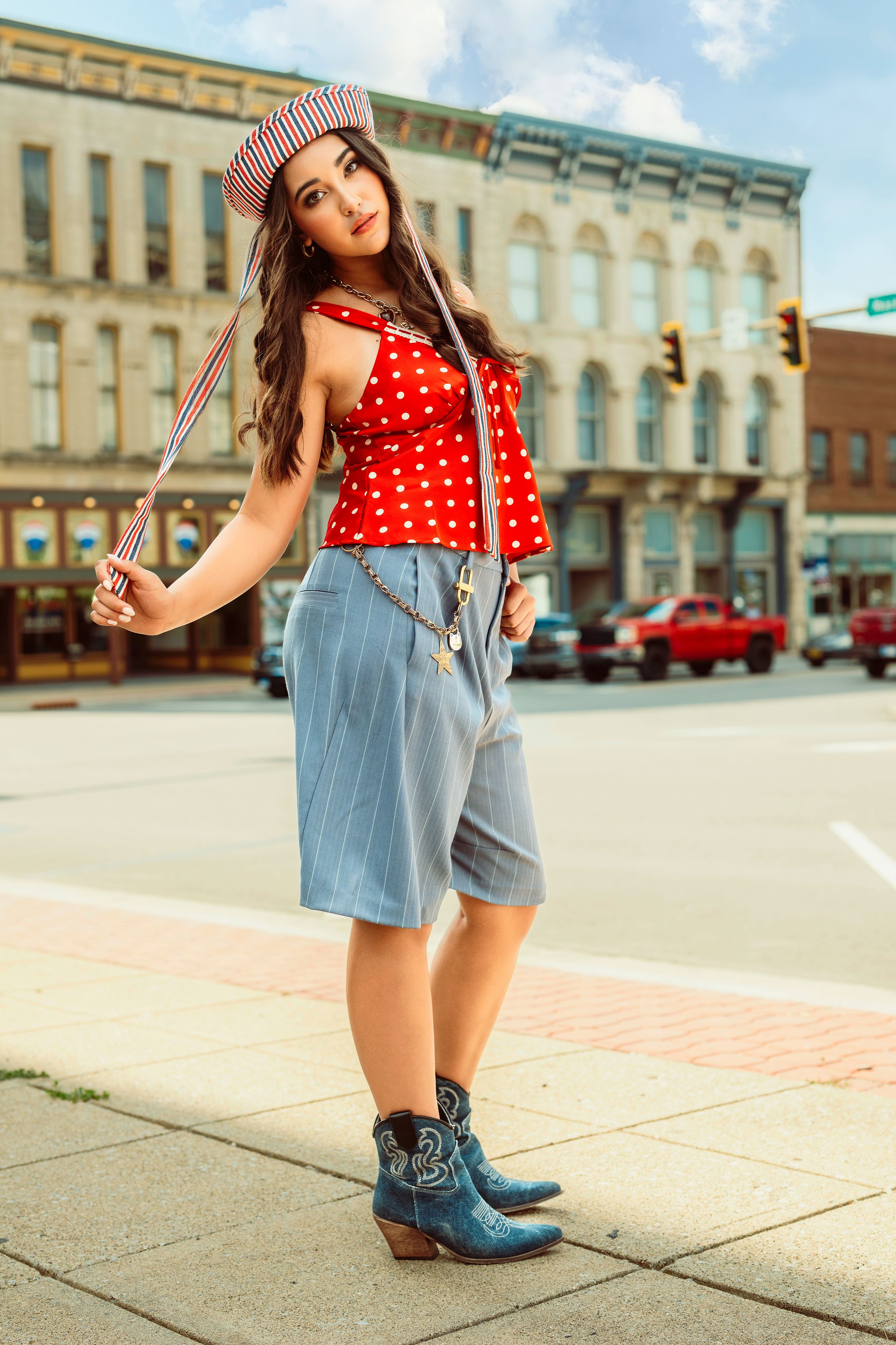 Stylish woman in red polka dot top, denim shorts, suspenders, and cowboy boots posing on a city sidewalk. Urban editorial