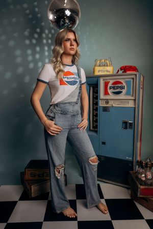 Blonde woman in Pepsi t-shirt and ripped flare jeans standing confidently near vintage Pepsi vending machine.