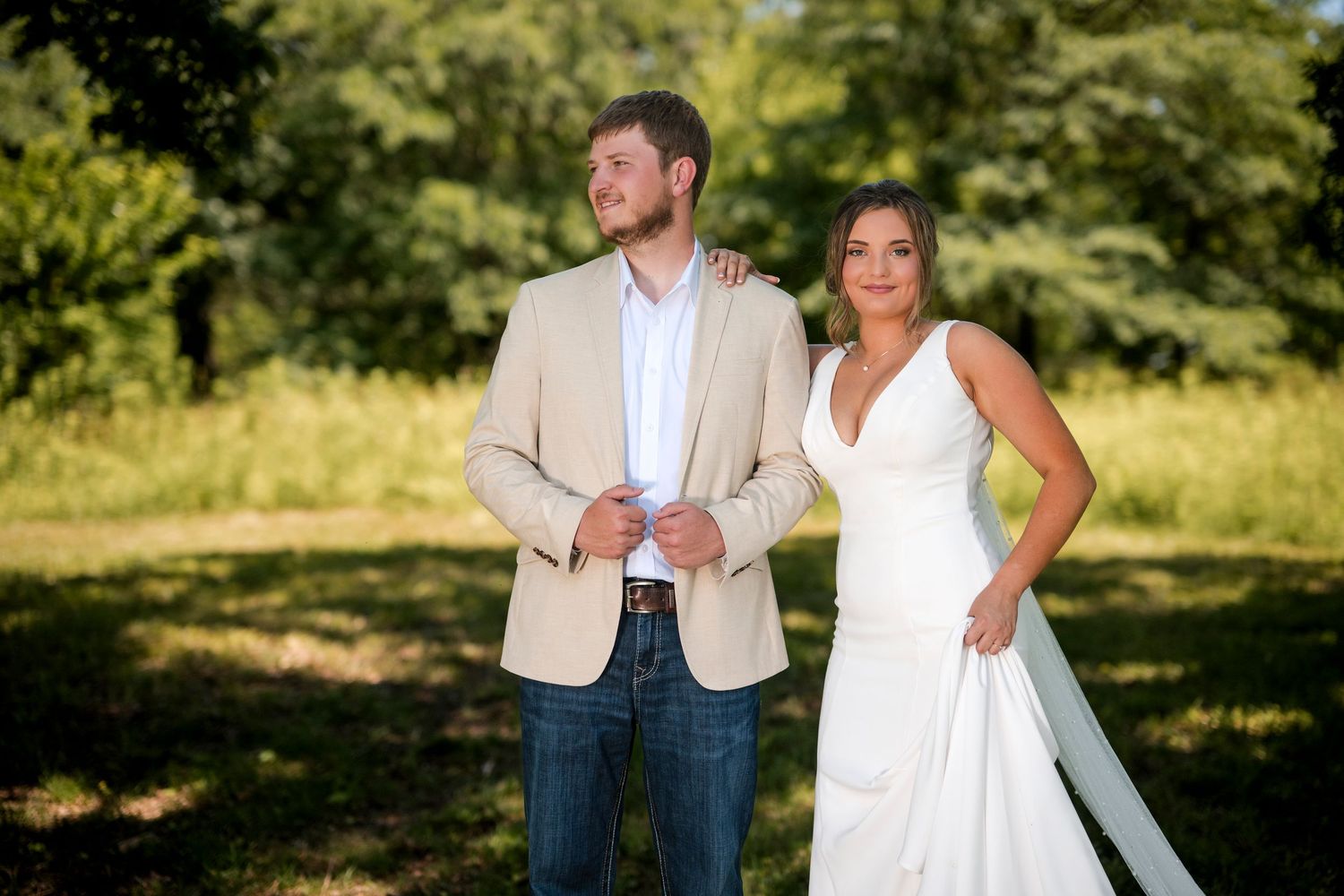 Bride in white wedding gown standing with groom in tan blazer and jeans in lush green outdoor setting.