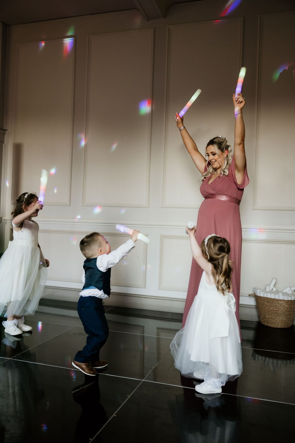 A bridesmaid in a pink gown dances with children holding glowing light sticks at a wedding reception.