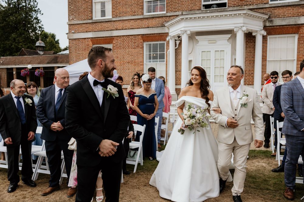 Bride walking down outdoor aisle with older gentleman, groom waiting ahead, guests seated at garden wedding.