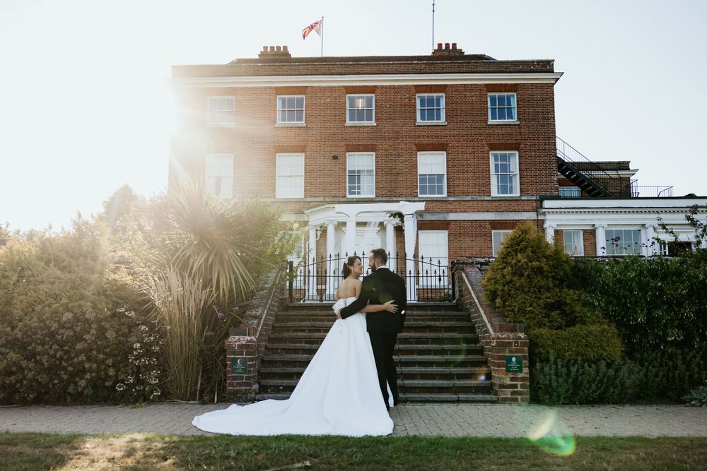 Bride and groom embrace in front of a grand red brick wedding venue with white columns and sunlit gardens.