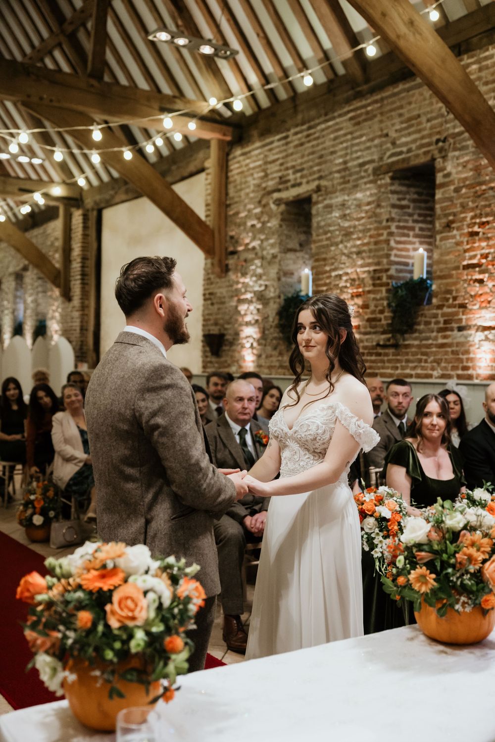 Couple exchanging vows in a rustic barn wedding venue with exposed brick walls, wooden beams, and orange floral arrangements.
