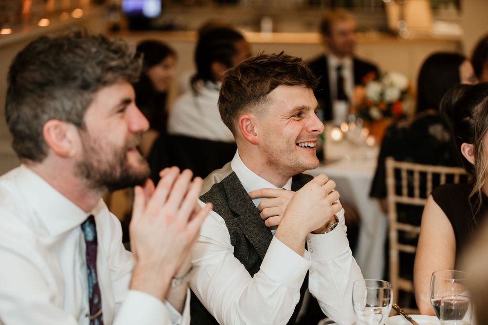 Two men in formal attire laugh and clap at a wedding reception dinner, one wearing a vest and tie.