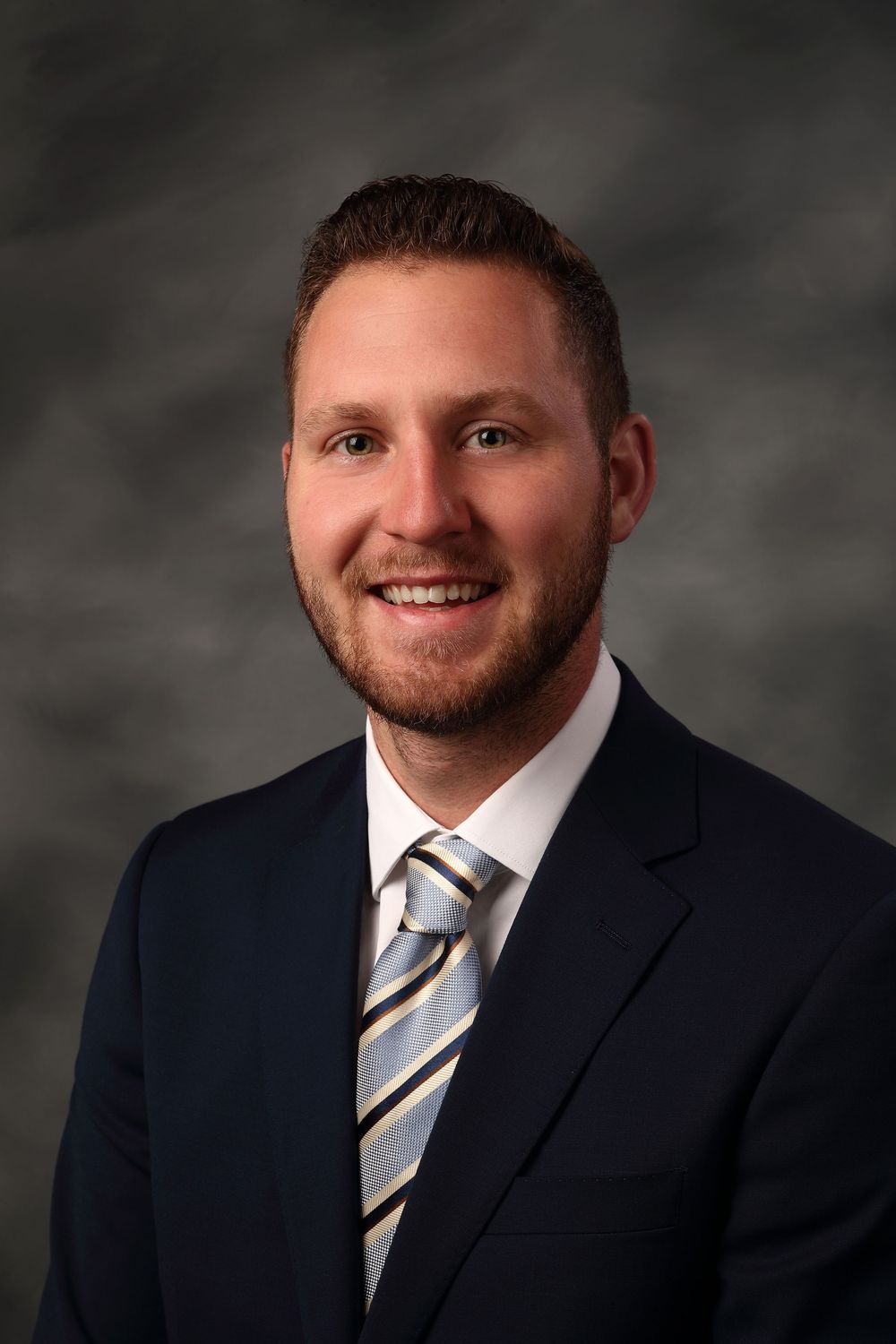 Professional headshot of someone in a dark suit and striped tie against a gray studio backdrop, smiling warmly at camera.