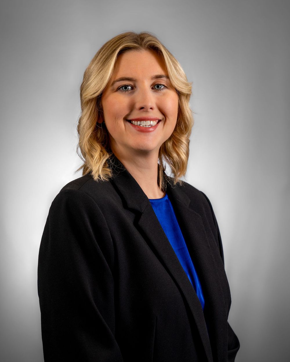 Professional headshot portrait of a person in a black blazer and blue top against a gray gradient background.