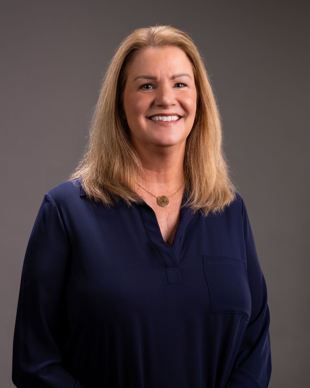 Professional headshot of a smiling blonde woman in a navy blue blouse with a gold necklace on a gray background.