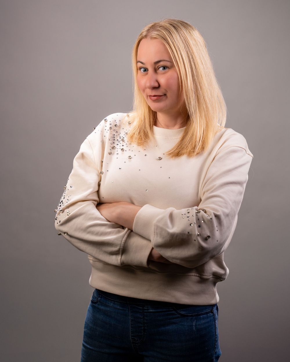 Blonde woman in beige embellished sweatshirt and jeans, arms crossed, posing confidently against gray background.