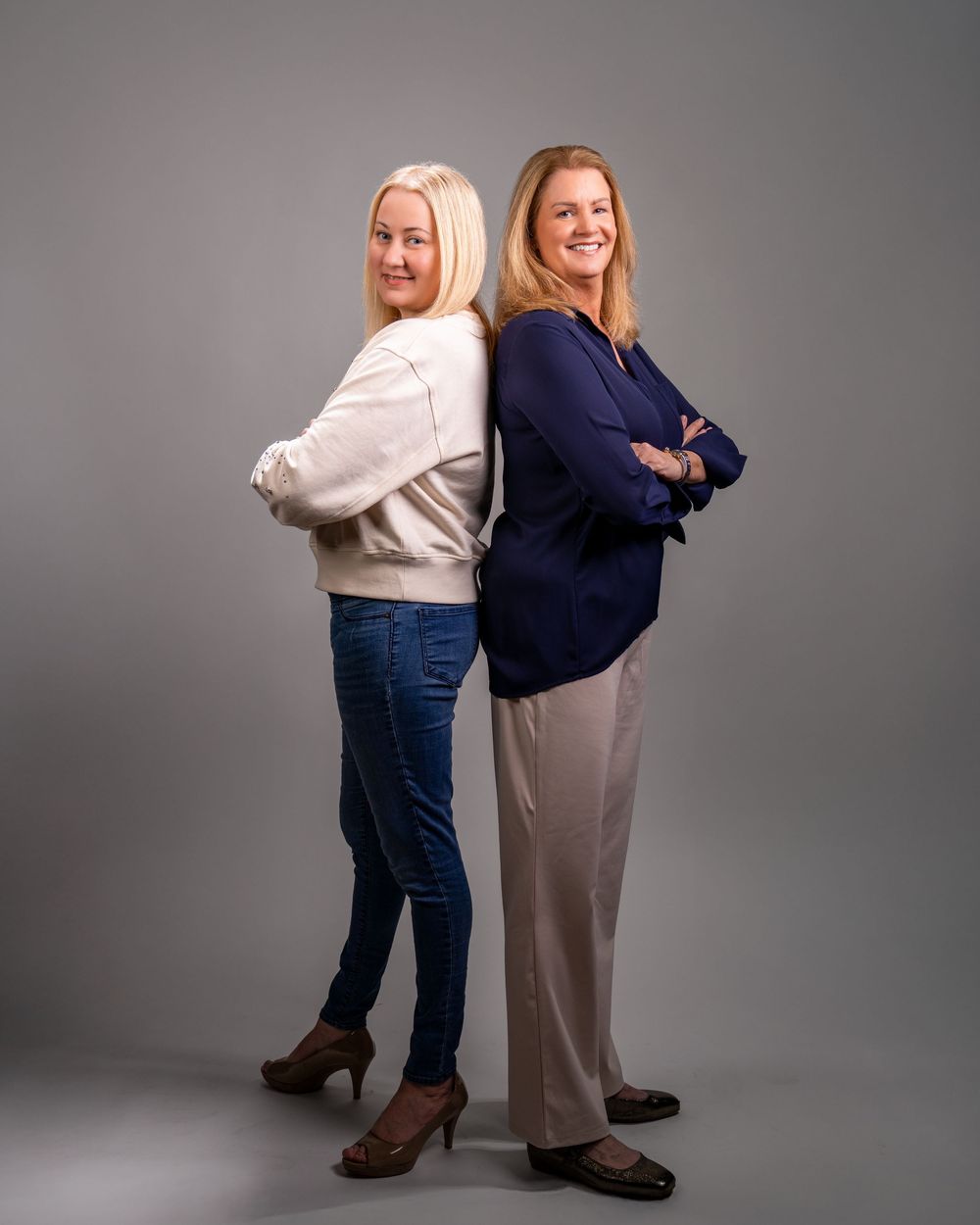 Two smiling women standing back-to-back with arms crossed in a professional studio portrait photo.