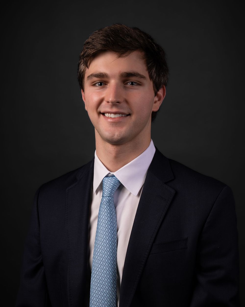 Professional headshot of a person in a navy suit and light blue tie smiling at camera against dark background.