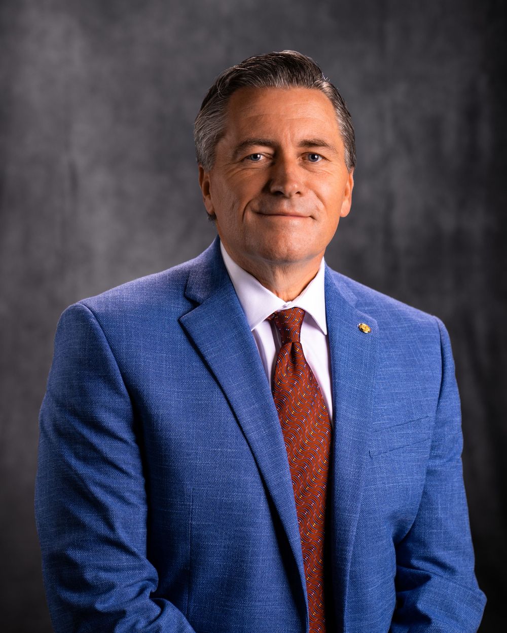 Professional headshot of person in blue suit and red tie against textured gray background.