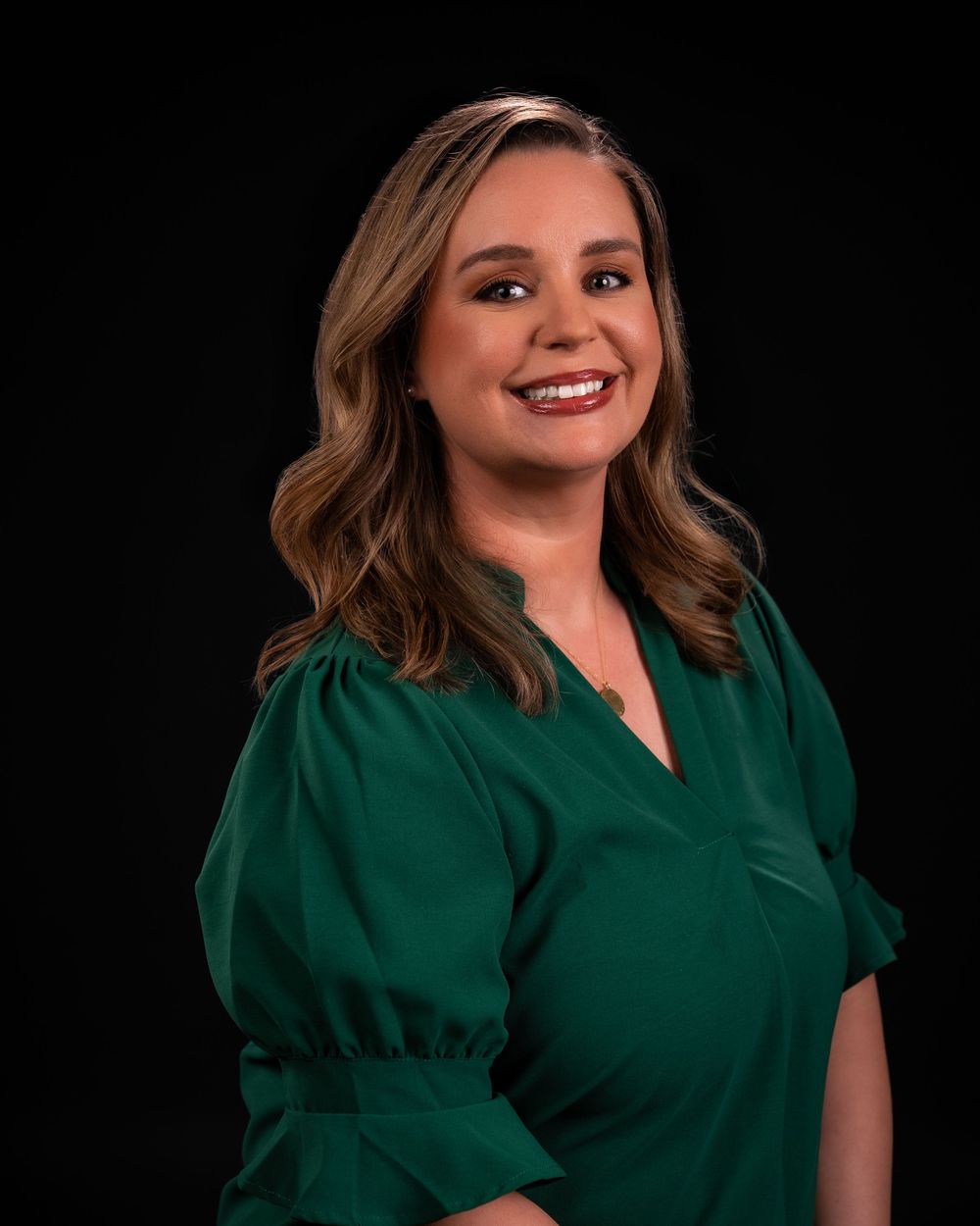 Professional headshot of a person wearing an emerald green v-neck top with puff sleeves against a black background.