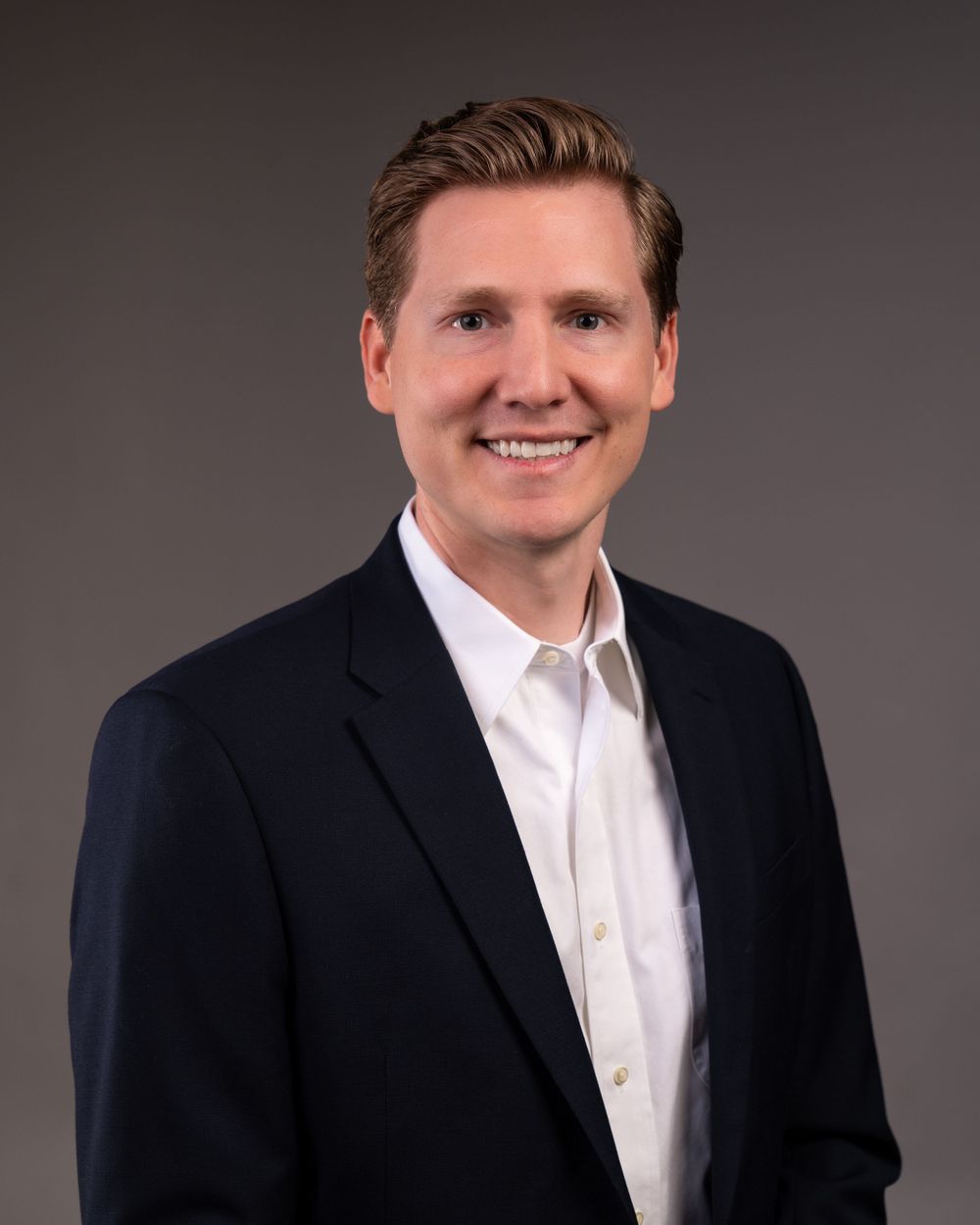 Professional headshot of person in navy suit jacket and white button-up shirt against dark background.